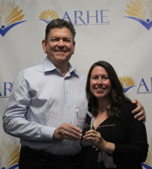 A smiling man and woman pose together holding a clear glass award in front of a backdrop with the ARHE logo and yellow-blue graphics.