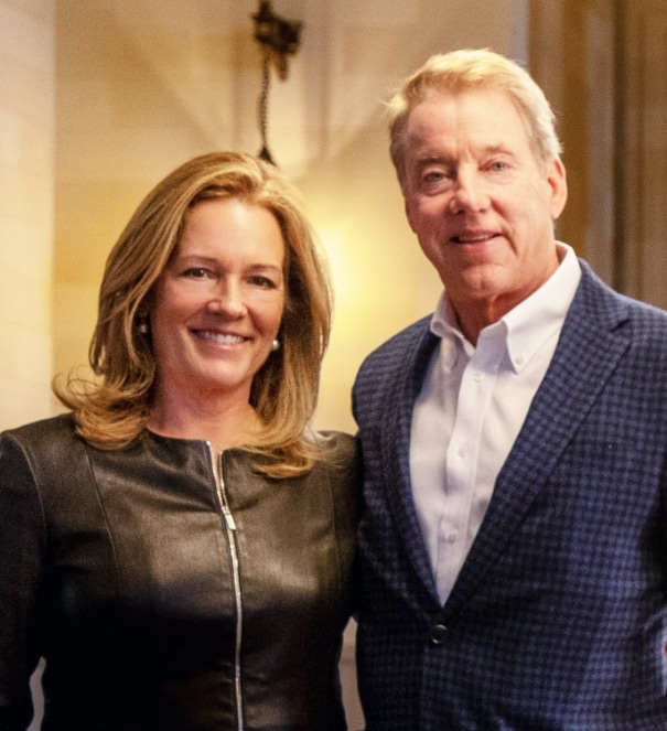 A woman and a man stand close together indoors, both smiling. The woman has shoulder-length blonde hair and wears a black leather jacket, while the man has light hair and wears a blue checked suit jacket over a white shirt.