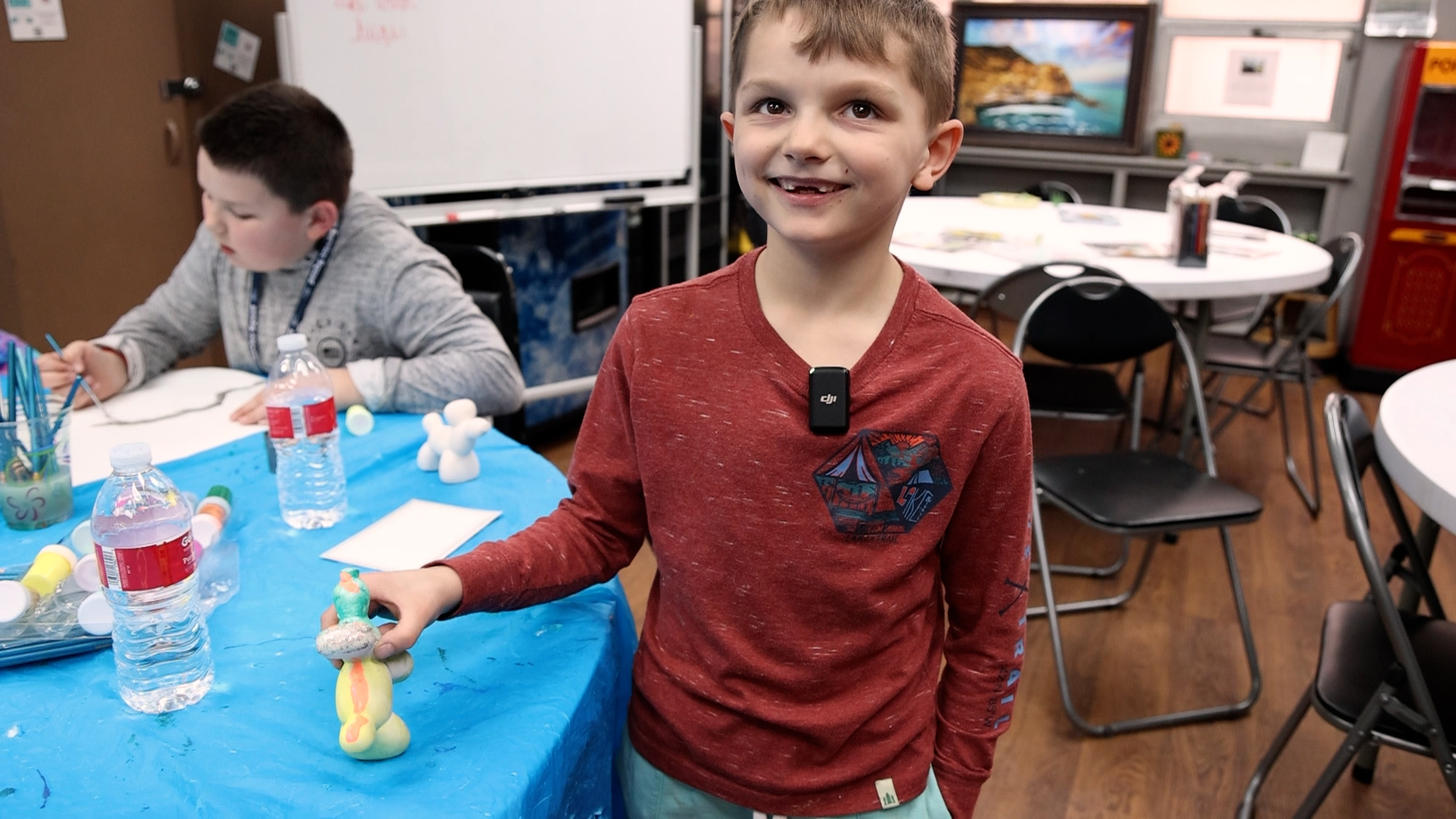 A smiling young boy stands next to a blue table, holding a colorful painted ceramic figure. Another child sits at the table painting. Art supplies and water bottles are visible, with a classroom in the background.
