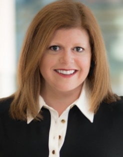 A woman with straight, shoulder-length light brown hair smiles at the camera. She is wearing a black top with a white collared shirt underneath. The background is softly blurred.