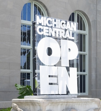 A large sign in front of a building reads "MICHIGAN CENTRAL OPEN" in bold white letters, with arched windows and stone walls visible in the background.