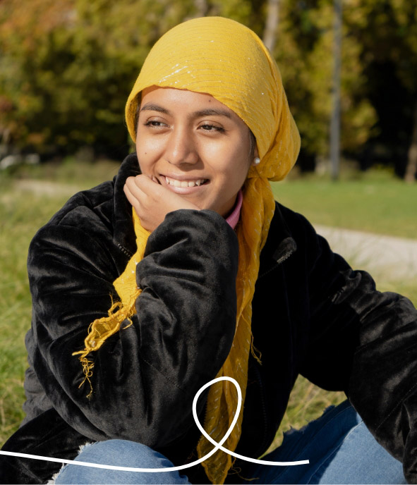 A young person in a yellow headscarf and black jacket sits outdoors on grass, smiling and looking to the side. Trees and sunlight are visible in the background, with a white abstract line at the bottom of the image.
