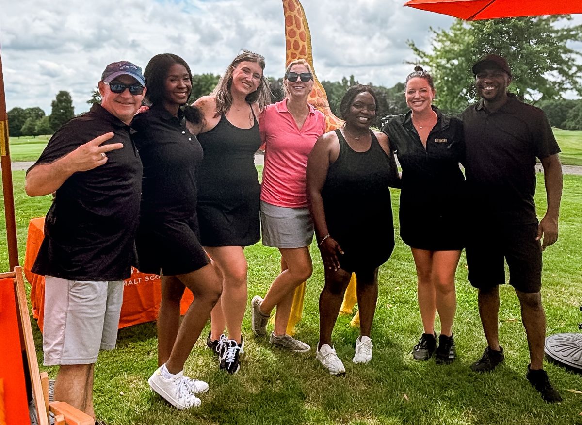 A group of seven people, dressed in golf attire, stand smiling together outdoors on a grassy area during daytime. Some wear black, one wears pink, and there is a giraffe-patterned flag behind them.