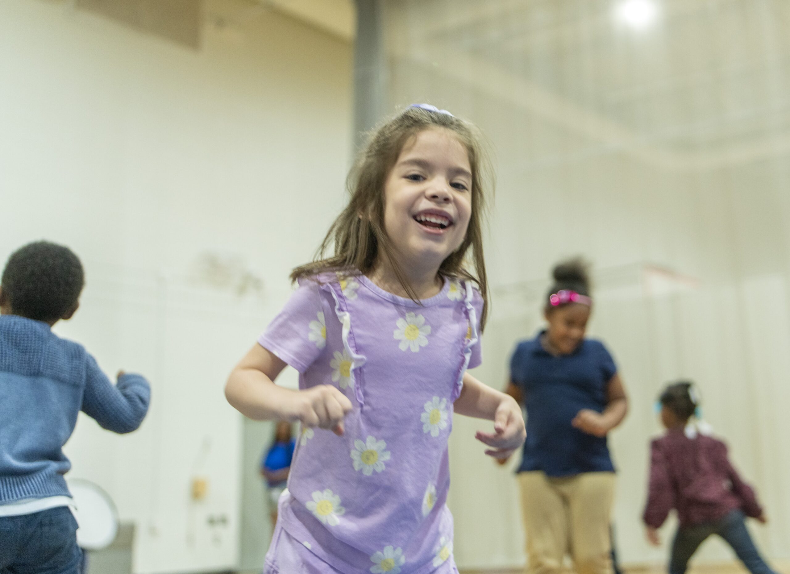 A young girl in a purple shirt with white daisies smiles and plays indoors with other children in the background. The setting appears to be a gym or recreational area.
