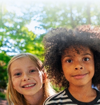 Two children, one with long blonde hair and one with curly dark hair, smile and stand close together outdoors with green trees and sunlight in the background.