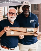 Two smiling men in navy blue shirts pose together. One holds a wooden plaque with a mounted cane and nameplate. They are standing outdoors under a canopy.