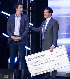 Two men in suits stand on a stage; one smiles while holding an oversized check for $27,500 made out to a foundation. Stage lights and blue accents are visible in the background.