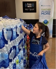A young girl wearing a blue apron reaches for a water bottle from a large stacked display of bottled water, with a “free WiFi Hot Spot” sign and a community banner in the background.