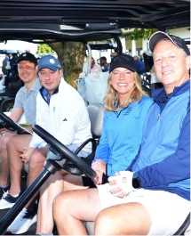 Four adults sit together in a golf cart, smiling at the camera. They are dressed in casual golf attire, including caps and jackets, with a golf course visible in the background.