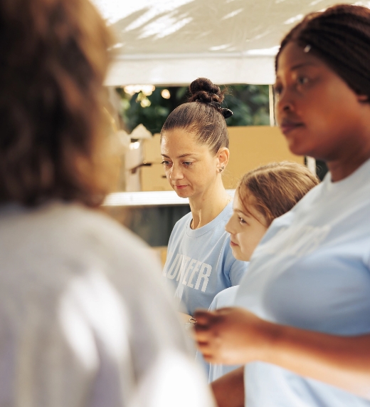 Three women and a young girl wearing light blue shirts stand together outdoors, appearing to be at a volunteer event. The focus is on the two women and girl in the background, with soft sunlight illuminating the scene.