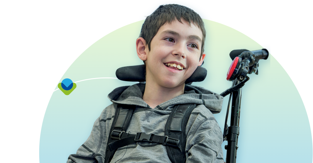 Smiling boy with short dark hair wearing a gray hoodie sits in a wheelchair with a supportive headrest against a light gradient background.