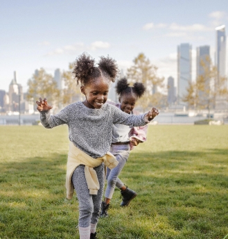 Two young girls with pigtails play and run on green grass in a city park, with tall buildings and a blue sky in the background. Both are smiling and wearing casual clothes.