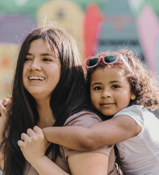 A smiling woman with long dark hair stands outdoors as a young girl with curly hair and pink sunglasses on her head hugs her from behind, both looking happy. Colorful shapes are blurred in the background.