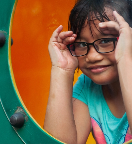 A young child with glasses smiles and looks through a circular window on a playground. The background is orange, and the child is wearing a blue shirt with a butterfly design.