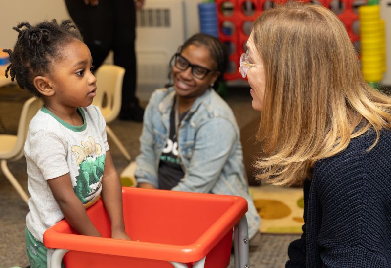 A young child stands in a red cart, looking at an adult woman with glasses who is smiling and talking to them, while another woman sits on the floor in the background, smiling.