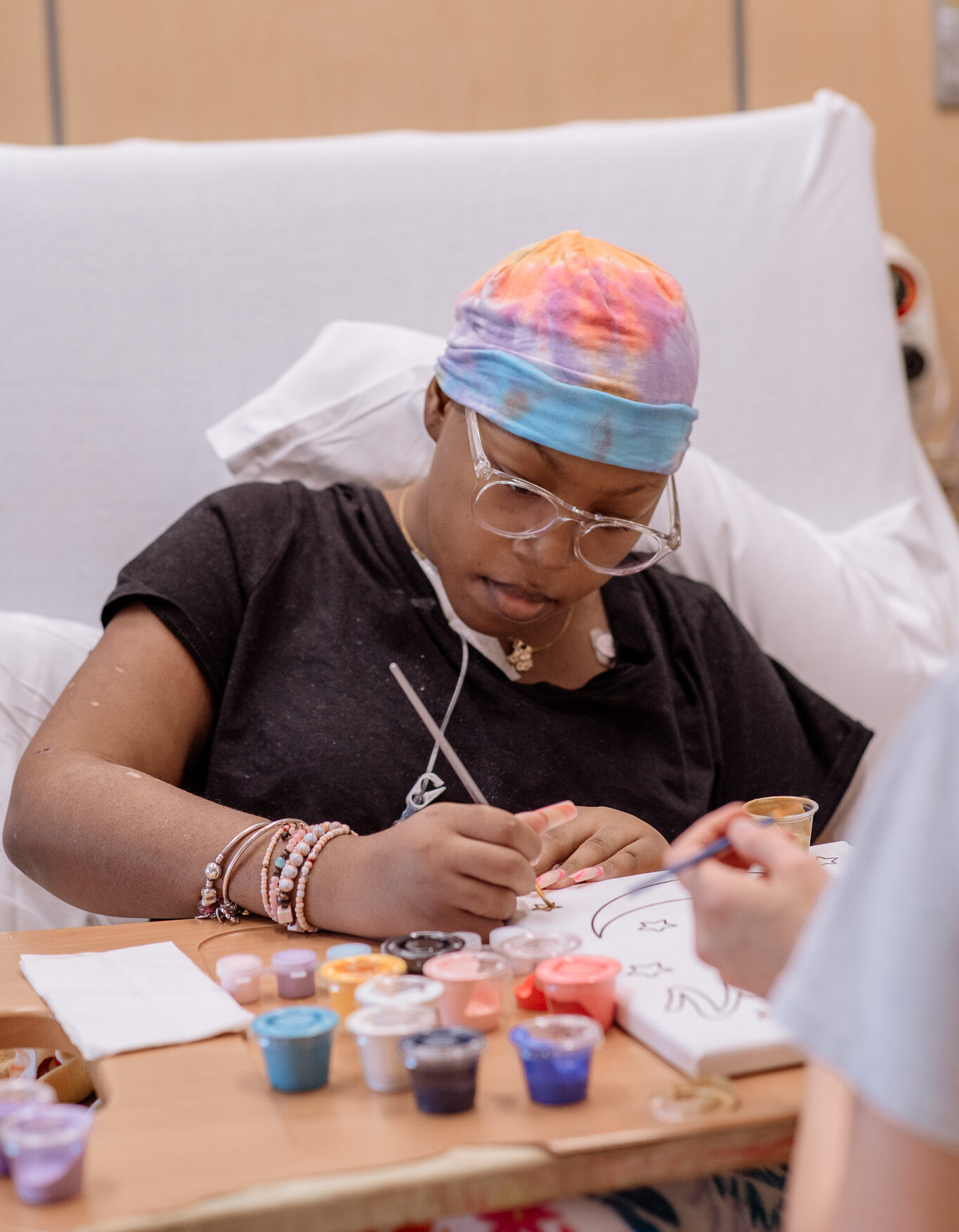 A person wearing a colorful headscarf and glasses sits in a hospital bed, focused on painting a design on paper. Various paint pots and art supplies are arranged on a tray in front of them.