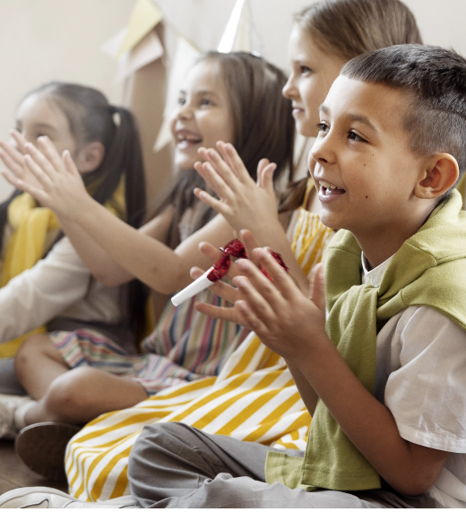 Four young children sit side by side on the floor, smiling and clapping their hands. They appear to be engaged in an activity, wearing light, colorful clothing and looking cheerful and attentive.