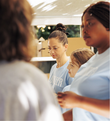 A group of women and a girl, all wearing light blue shirts, stand together outdoors. One woman is in focus, looking down thoughtfully, while others are slightly blurred in the foreground.
