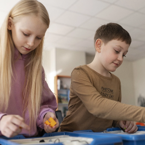 Two children are building with small plastic blocks at a table in a classroom. The girl on the left wears a pink hoodie, and the boy on the right wears a brown shirt. They are focused on their activity.