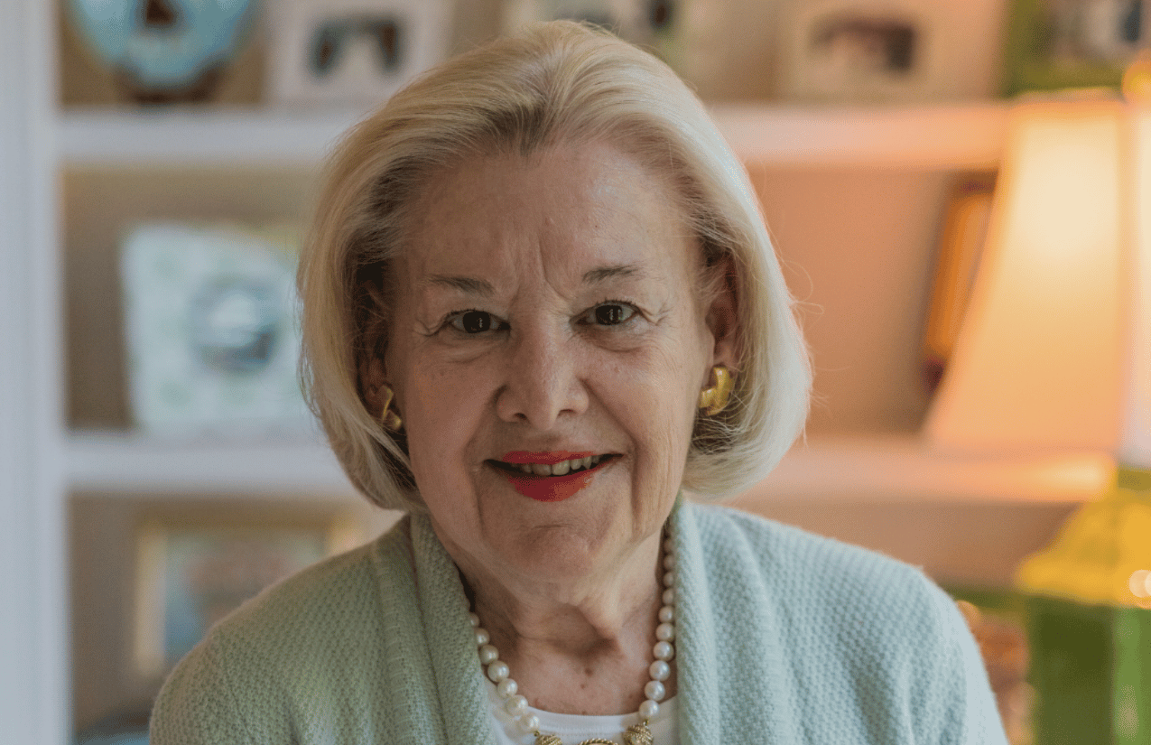 An older woman with light blonde hair and red lipstick, wearing a light green sweater, pearl necklace, and gold earrings, smiles at the camera. She is indoors with softly lit shelves and a lamp in the background.