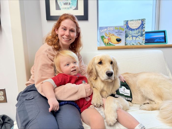 A woman and a young child sit on a couch next to a golden retriever wearing a service vest. The child is on the woman's lap and the dog rests its head on the child. Books are visible on the windowsill behind them.