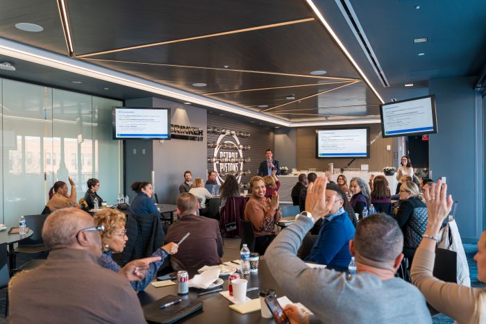People seated around tables in a modern conference room listen to a speaker at the front. Several attendees have their hands raised, and presentation slides are displayed on large screens.