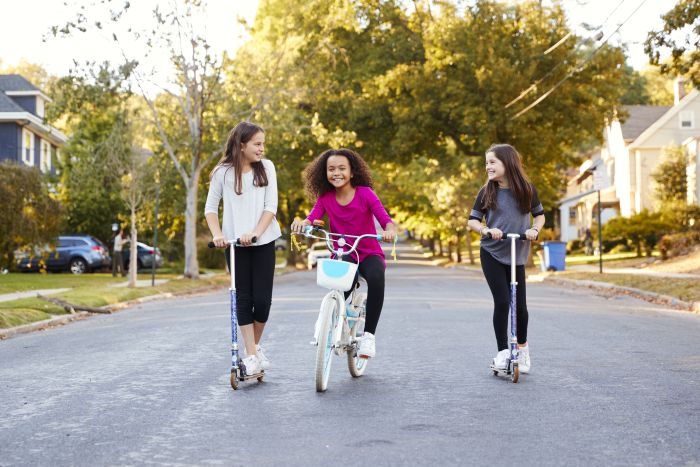 Three young girls play on a suburban street; two ride scooters while the one in the middle rides a bicycle. They are smiling and interacting with each other, surrounded by houses and trees on a sunny day.