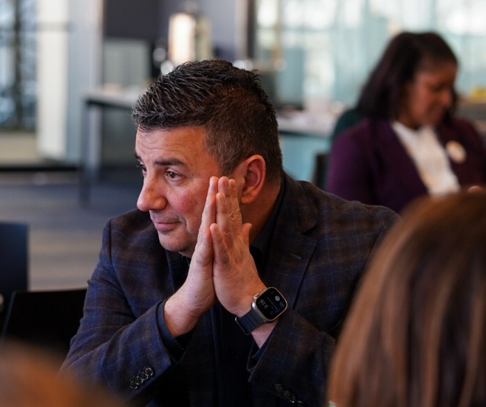 A man in a plaid jacket sits at a table with his hands pressed together, appearing thoughtful. He wears a smartwatch. In the background, a woman sits and looks downward. The setting appears to be a modern indoor office or meeting space.