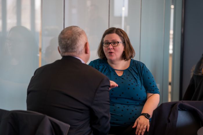 A woman in a blue polka dot shirt and glasses is talking to a man in a suit. They are seated indoors, engaged in conversation in a modern office setting with glass walls.