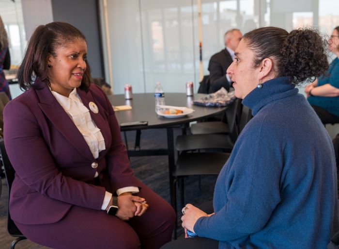 Two women sit facing each other and talking in a professional setting. One wears a maroon suit and the other a dark blue sweater. Other people are seen conversing in the background at tables.