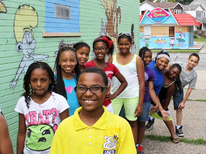 A group of smiling children stand outside in front of a colorful mural wall and an ice cream shop, posing for the camera on a sunny day.