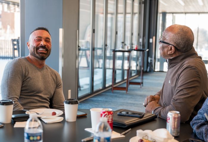 Two men sit at a table, smiling and laughing together. The table has coffee cups, water bottles, and soda cans. Large windows in the background let in natural light, creating a casual and friendly atmosphere.