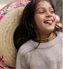 A young girl with long brown hair smiles playfully with her tongue out, wearing a light sweater and a large woven straw hat with colorful trim resting on her back.