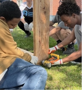 Two people attach a wooden beam to a post outdoors using a power drill, while others sit nearby helping with the construction project. Grass and a brick building are visible in the background.