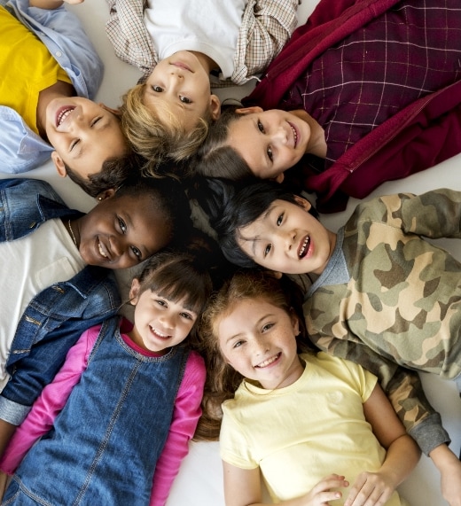 Seven children of diverse backgrounds lie on the floor in a circle with their heads together, smiling up at the camera. They wear colorful, casual clothes and appear happy and cheerful.