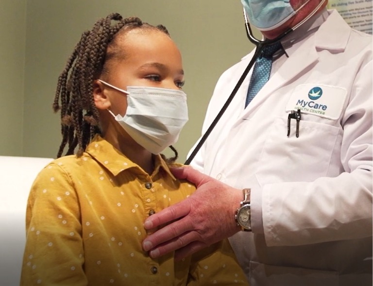 A young girl wearing a face mask and yellow shirt sits while a doctor with a stethoscope, also masked and in a white coat, examines her at a medical clinic.