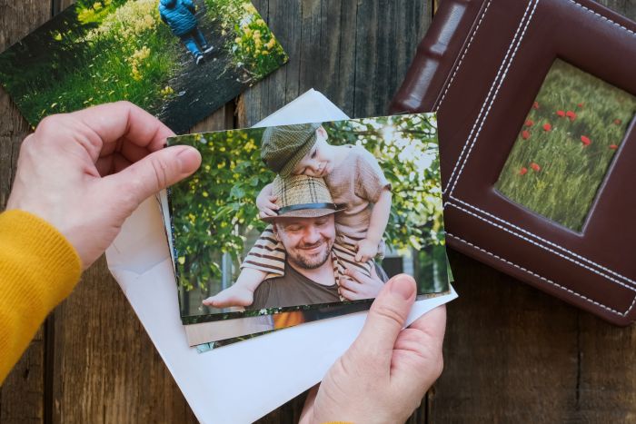 A person holds a photo of a man with a child on his shoulders, both smiling outdoors. An envelope, another photo, and a leather photo album are on a rustic wooden table.