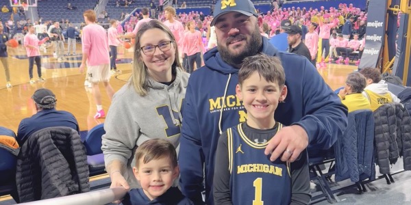 A smiling family of four in Michigan gear poses at a basketball game. The court and players in pink shirts are visible in the background, along with spectators in the stands.