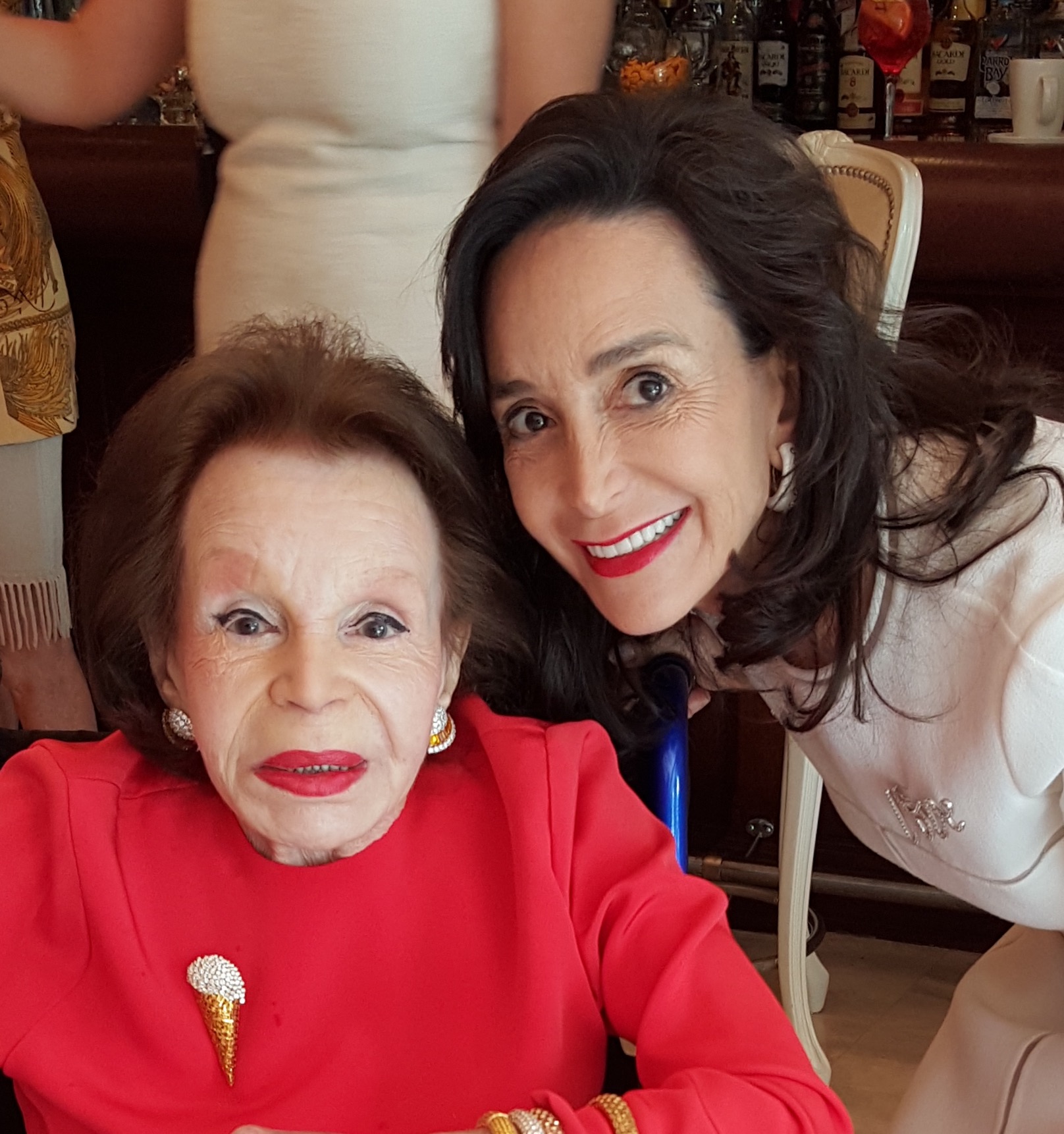 Two women pose closely together and smile. The older woman, in a jeweled red top and gold jewelry, shares a warm moment with the younger woman in white. Their bond reflects the spirit of legacy giving. A bar with bottles is visible in the background.